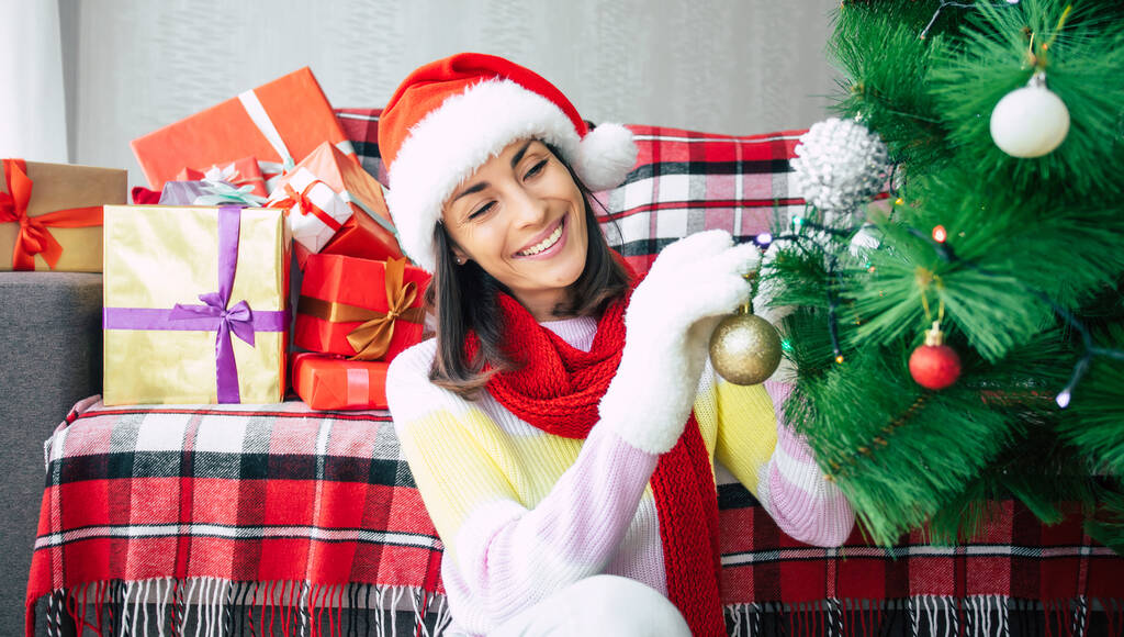 Foto de cerca de una hermosa mujer joven feliz en un suéter y sombrero de santa está decorando un árbol de Navidad en casa en el fondo de las cajas de regalo. Vacaciones de invierno - Foto, Imagen