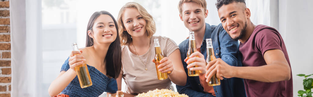 panoramisch concept van vreugdevolle multi-etnische vrienden die naar de camera kijken terwijl ze flessen bier vasthouden  - Foto, afbeelding