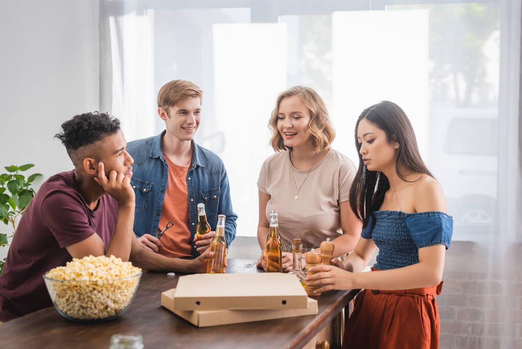 multiculturele vrienden aan het praten tijdens feest in de buurt van bier, popcorn en pizzadozen - Foto, afbeelding