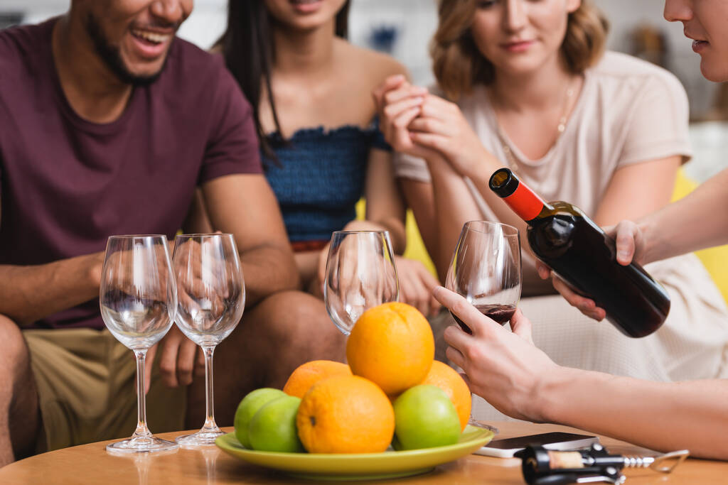 обрезанный вид молодого человека, разливающего красное вино рядом с фруктами и мультикультурными друзьями - Фото, изображение