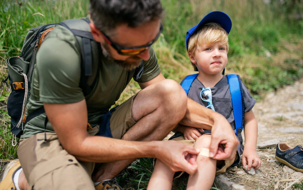 Father with small son hiking outdoors in summer nature, putting plaster on knee. - Photo, Image