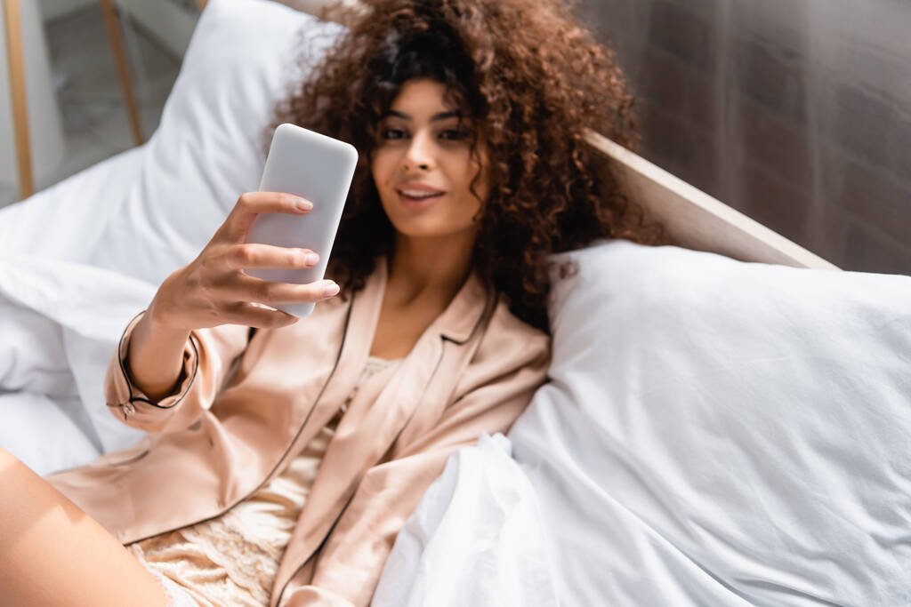 selective focus of curly woman taking selfie in bedroom - Photo, Image