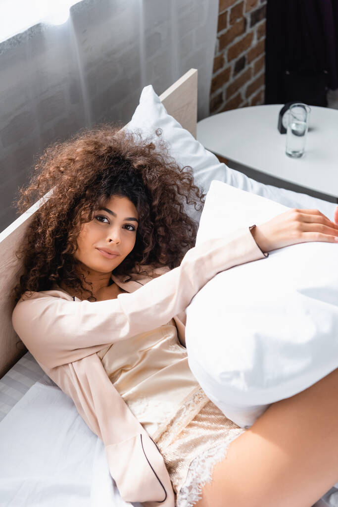 άποψη υψηλής γωνίας σγουρά νεαρή γυναίκα ξαπλωμένη στο κρεβάτι και κρατώντας μαξιλάρι - Φωτογραφία, εικόνα