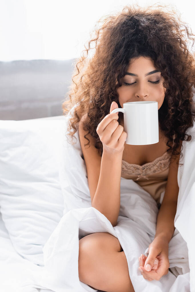  joven mujer rizada con los ojos cerrados oliendo café en el dormitorio - Foto, imagen