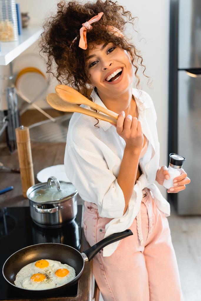 freudige Frau hält Küchenzange und Flasche mit Salz in der Nähe von Eiern in der Pfanne  - Foto, Bild