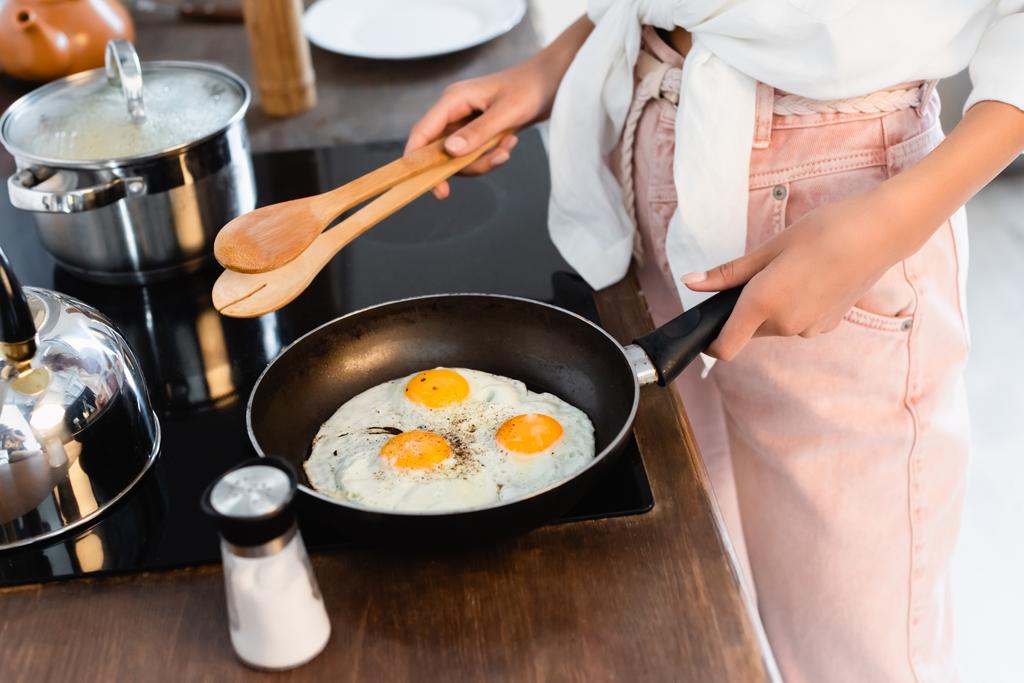 Ausgeschnittene Ansicht einer Frau, die eine Küchenzange in der Nähe von Eiern in der Pfanne hält - Foto, Bild
