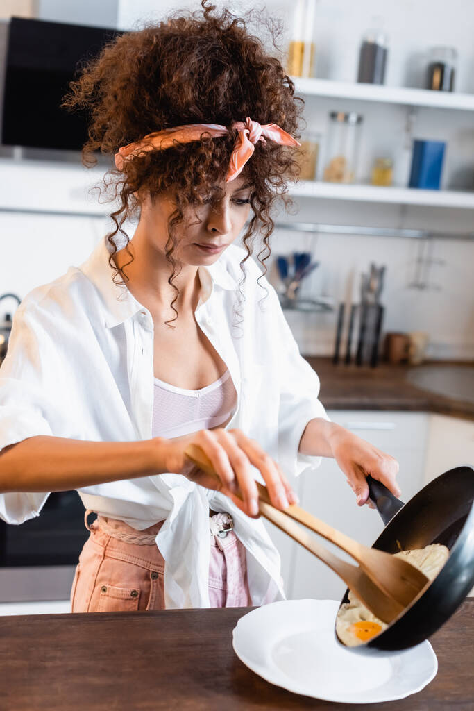 curly young woman holding frying pan and kitchen tongs while serving fried eggs on plate - Photo, Image