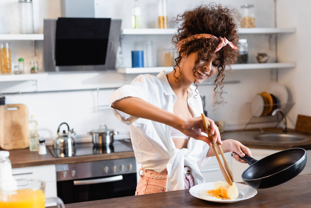 femme bouclée et joyeuse tenant poêle et pinces de cuisine tout en servant des œufs frits sur assiette - Photo, image