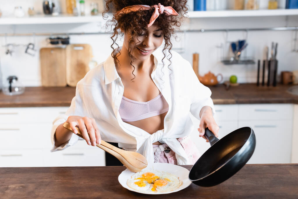 радісна жінка тримає сковороду і кухонні щипці, подаючи смажені яйця на тарілці
 - Фото, зображення
