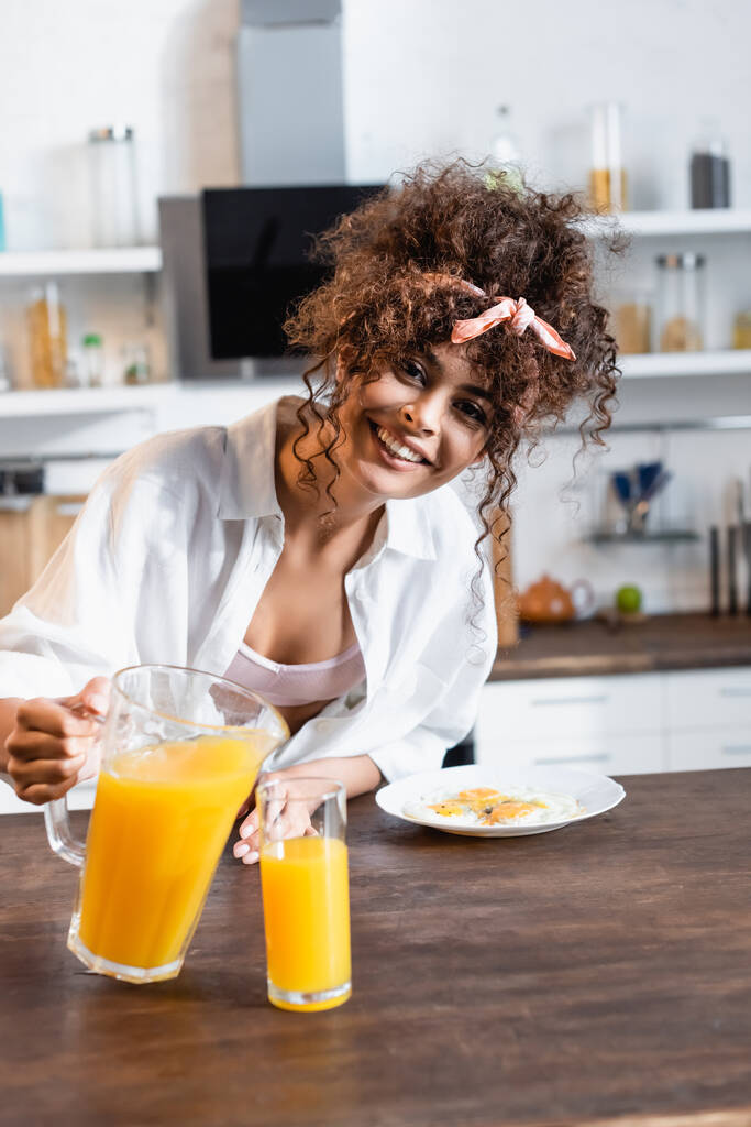 vreugdevolle vrouw houdt kruik in de buurt van glas en ontbijt op bord - Foto, afbeelding