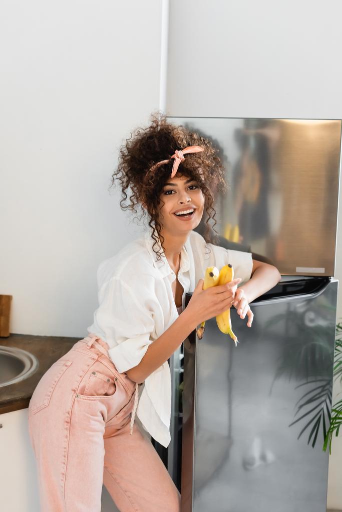 台所の冷蔵庫の近くでバナナを持っている楽しい女性  - 写真・画像