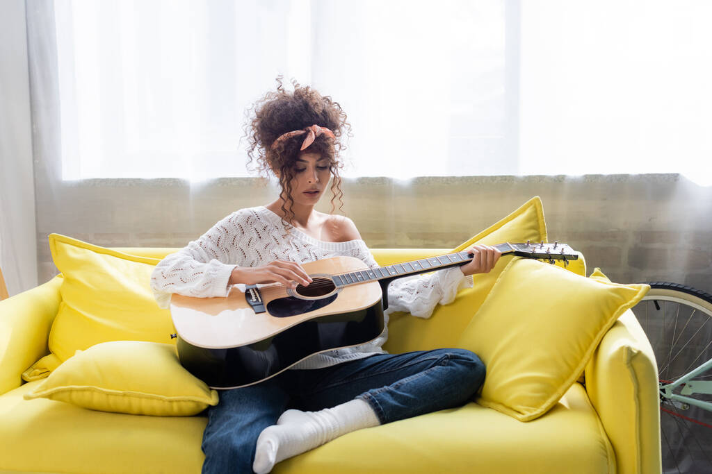 femme bouclée jouant de la guitare acoustique sur le canapé dans le salon  - Photo, image