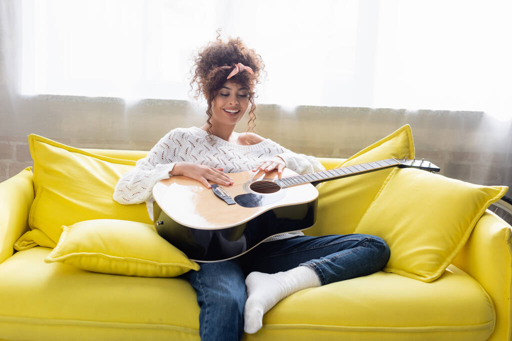 ευχαριστημένη γυναίκα που κρατάει ακουστική κιθάρα και κάθεται στον καναπέ στο σαλόνι  - Φωτογραφία, εικόνα