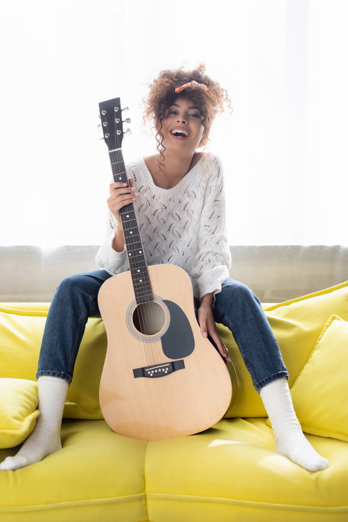 возбужденная женщина, держащая акустическую гитару и сидя на диване в гостиной  - Фото, изображение