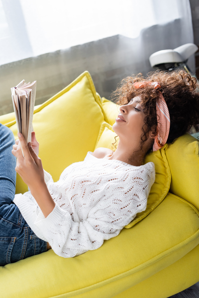 порадовала молодая женщина, читающая книгу лежа на диване в гостиной  - Фото, изображение