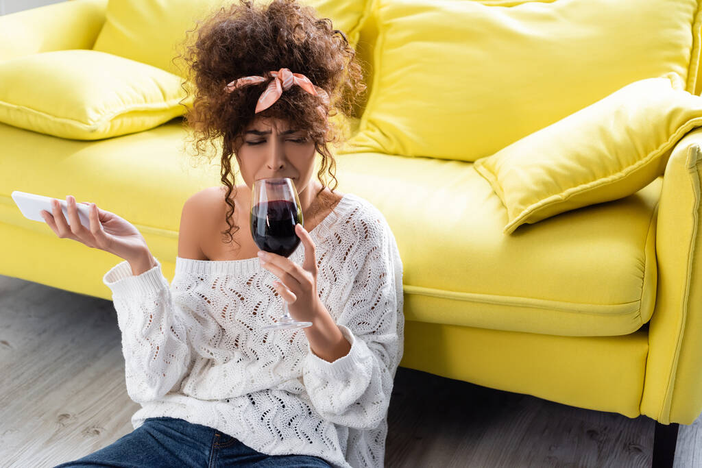 засмучена жінка тримає келих вина і смартфона під час плачу у вітальні
 - Фото, зображення
