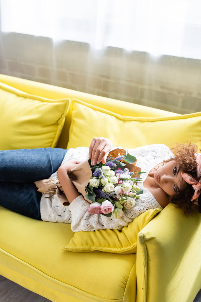 joven mujer rizada acostada en un sofá amarillo con flores y mirando hacia otro lado - Foto, Imagen