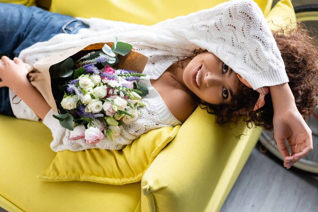 Overhead-Ansicht der fröhlichen und lockigen Frau mit Blumen auf der Couch liegend - Foto, Bild