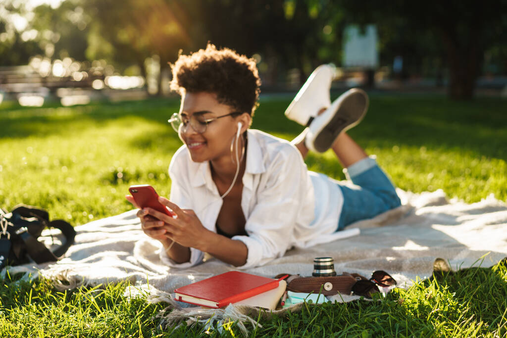 Photo de femme africaine optimiste heureuse couchée à l'extérieur dans un parc sur une herbe et utilisant un téléphone portable tout en écoutant de la musique - Photo, image
