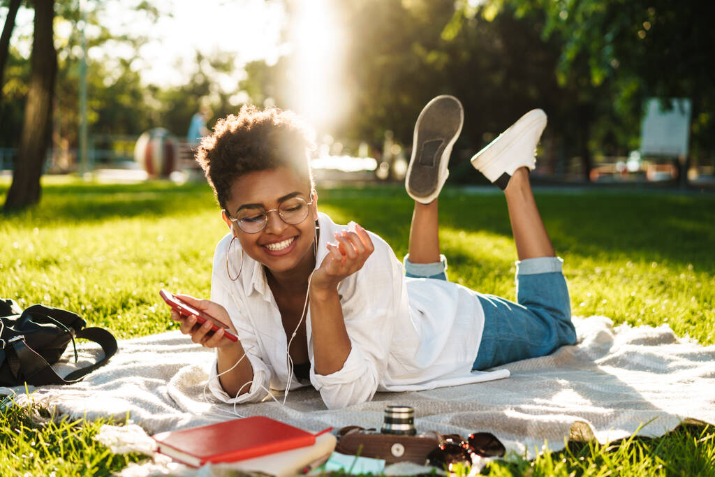 Zdjęcie uśmiechniętej afrykańskiej kobiety leżącej na zewnątrz w parku na trawie i korzystającej z telefonu komórkowego podczas słuchania muzyki - Zdjęcie, obraz