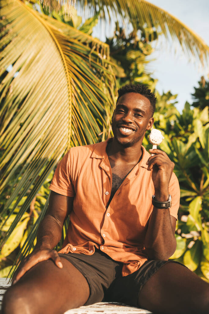 Fiatal, jóképű fekete férfi narancssárga inget visel, nyári napsütésben tartja és eszi a jégkrémet. - Fotó, kép