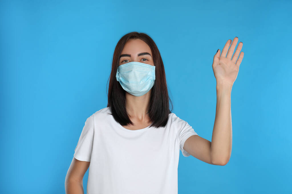 Junge Frau in Schutzmaske mit Hallo-Geste auf hellblauem Hintergrund. Soziale Distanz bei Coronavirus-Pandemie wahren - Foto, Bild