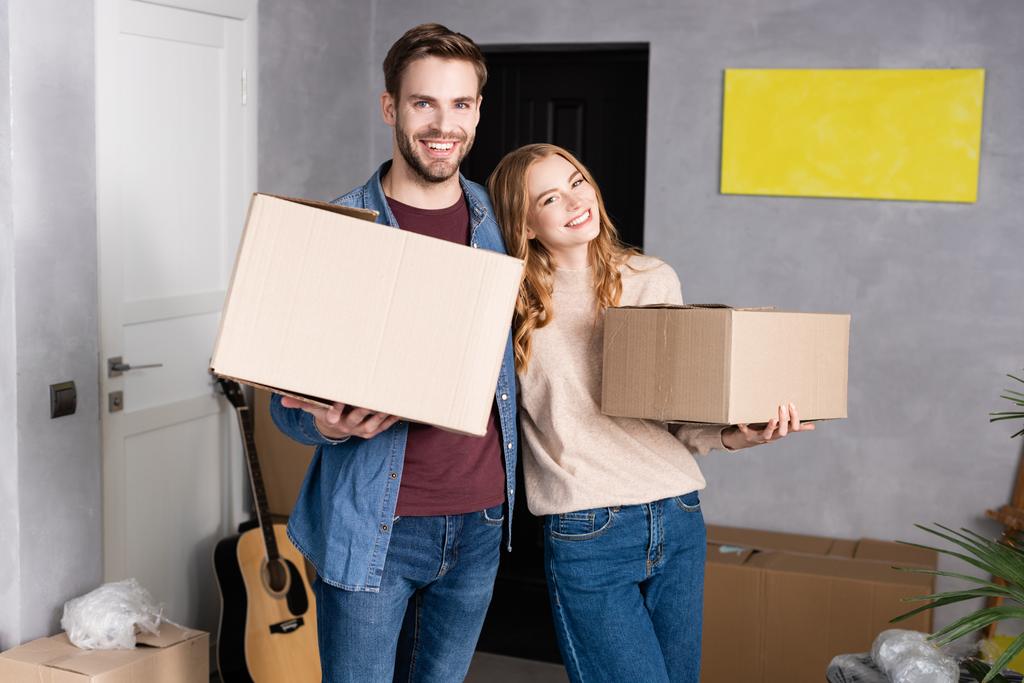 alegre hombre y mujer sosteniendo cajas de cartón en un nuevo hogar - Foto, imagen