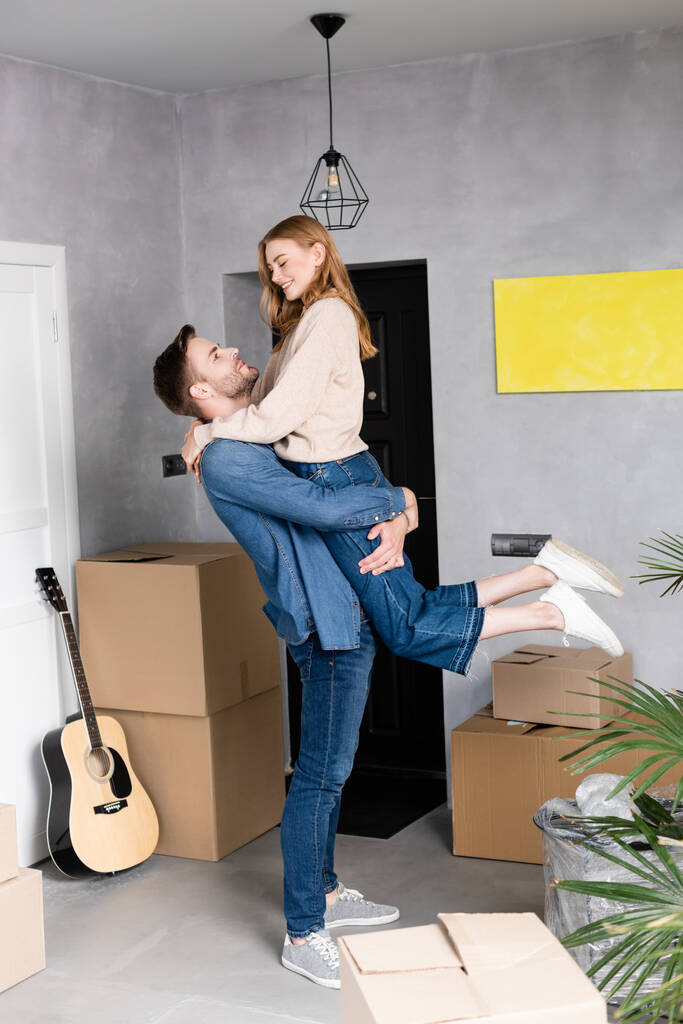 hombre sosteniendo en brazos complacido mujer cerca de cajas y guitarra acústica, concepto de reubicación - Foto, imagen