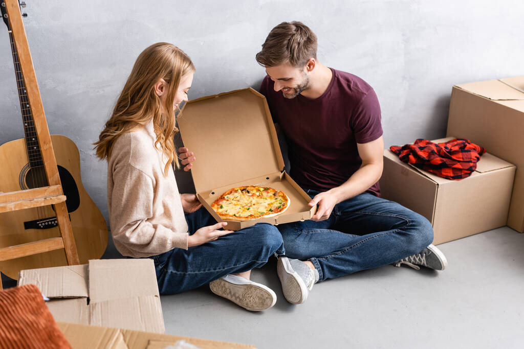 мужчина и женщина смотрят на пиццу в коробке рядом с пакетами и акустической гитарой - Фото, изображение