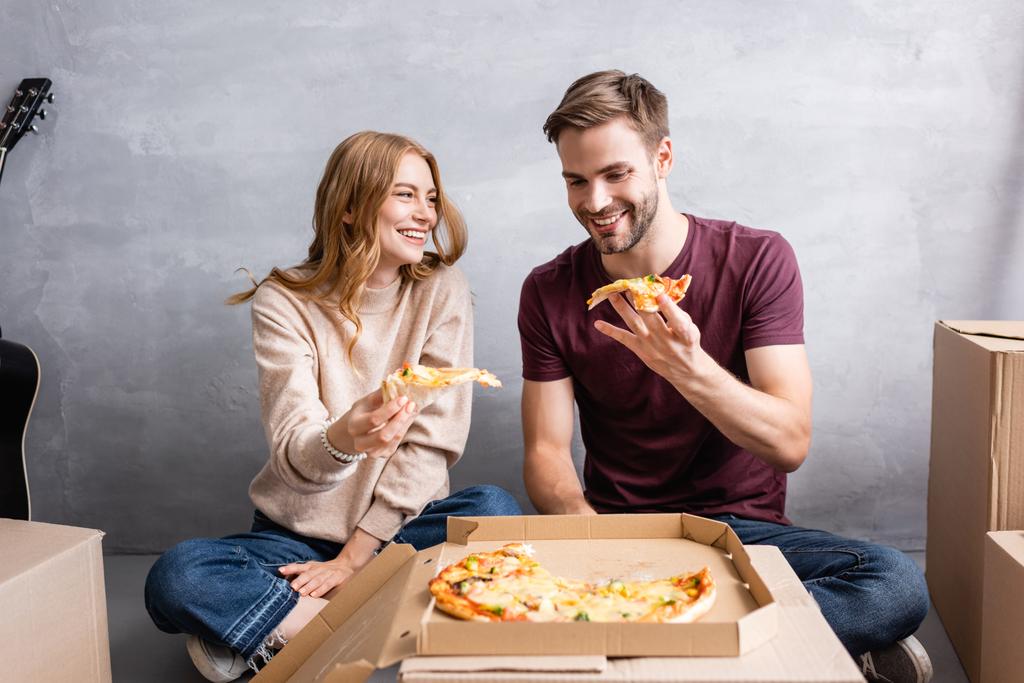 joyful woman looking at man while holding pizza in new home - Photo, Image