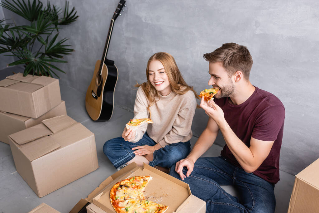 vista ad alto angolo di uomo seduto sul pavimento e mangiare pizza vicino a donna e scatole, concetto di trasferimento - Foto, immagini