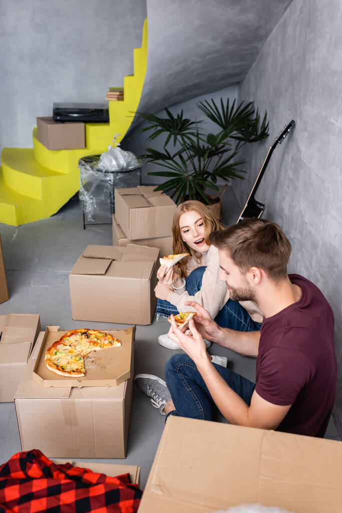 mulher com boca aberta olhando para o namorado com pizza nas mãos perto de caixas de papelão - Foto, Imagem