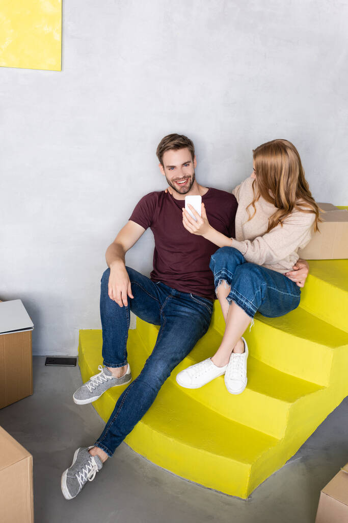 joyful man sitting on yellow stairs and using mobile phone near carton boxes and woman - Photo, Image