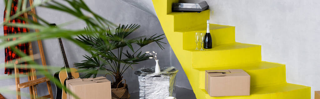 Panoramakonzept aus Kartons und Sektflasche auf gelben Treppen in der Nähe von Pflanzen und Akustikgitarre in moderner Wohnung  - Foto, Bild