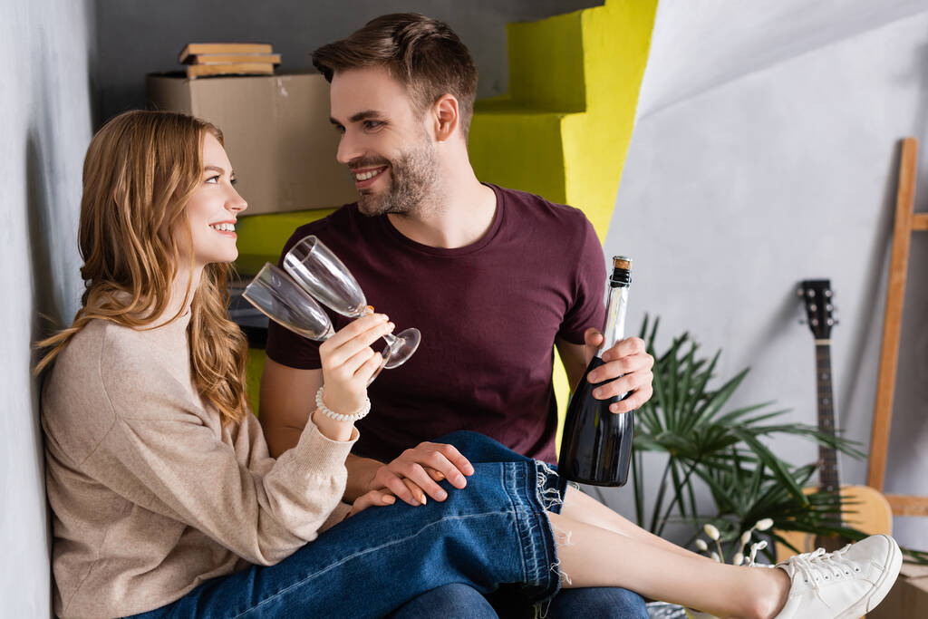 joyful couple holding bottle of champagne and glasses while looking at each other, moving concept - Photo, Image