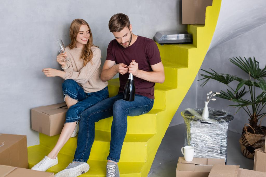 Mann öffnet Flasche, während junge Frau mit Brille auf gelber Treppe sitzt und sich bewegt  - Foto, Bild