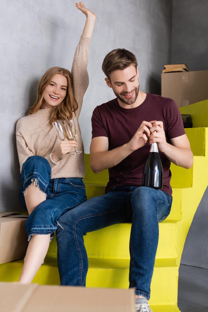 man opening bottle while pleased young woman holding glasses and sitting on stairs, moving concept  - Photo, Image