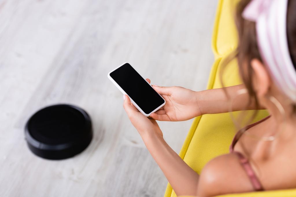 Enfoque selectivo de ama de casa utilizando el teléfono inteligente mientras está sentado en el sofá cerca de la aspiradora robótica  - Foto, imagen