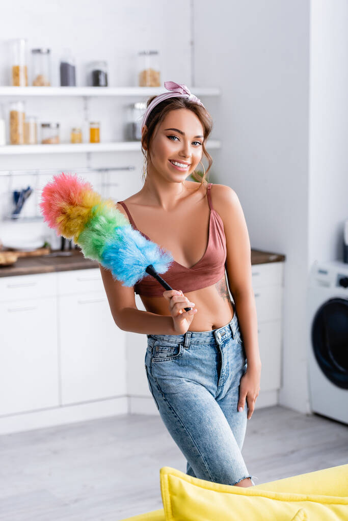 Selective focus of young housewife looking at camera and holding dust brush at home  - Photo, Image