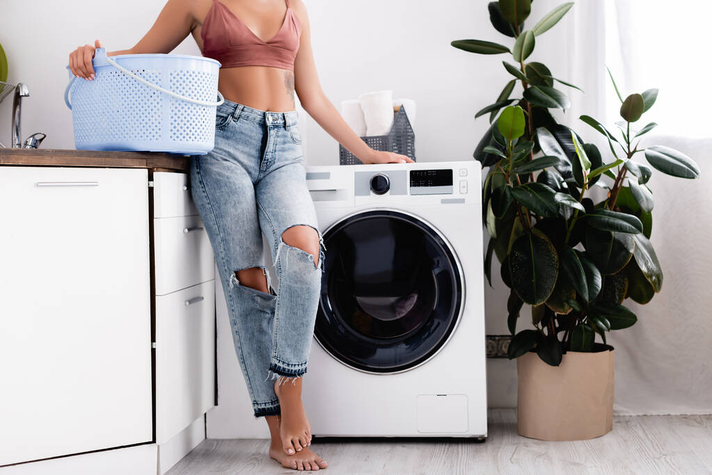 Vista recortada de una mujer descalza sosteniendo la cesta de la ropa cerca de la lavadora durante las tareas domésticas en casa  - Foto, Imagen