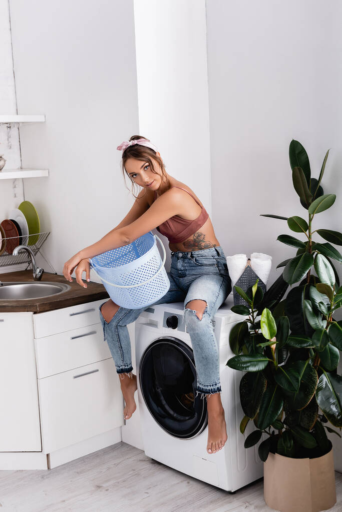 台所の洗濯機に座っている間に洗濯バスケットを持っている若い女性  - 写真・画像