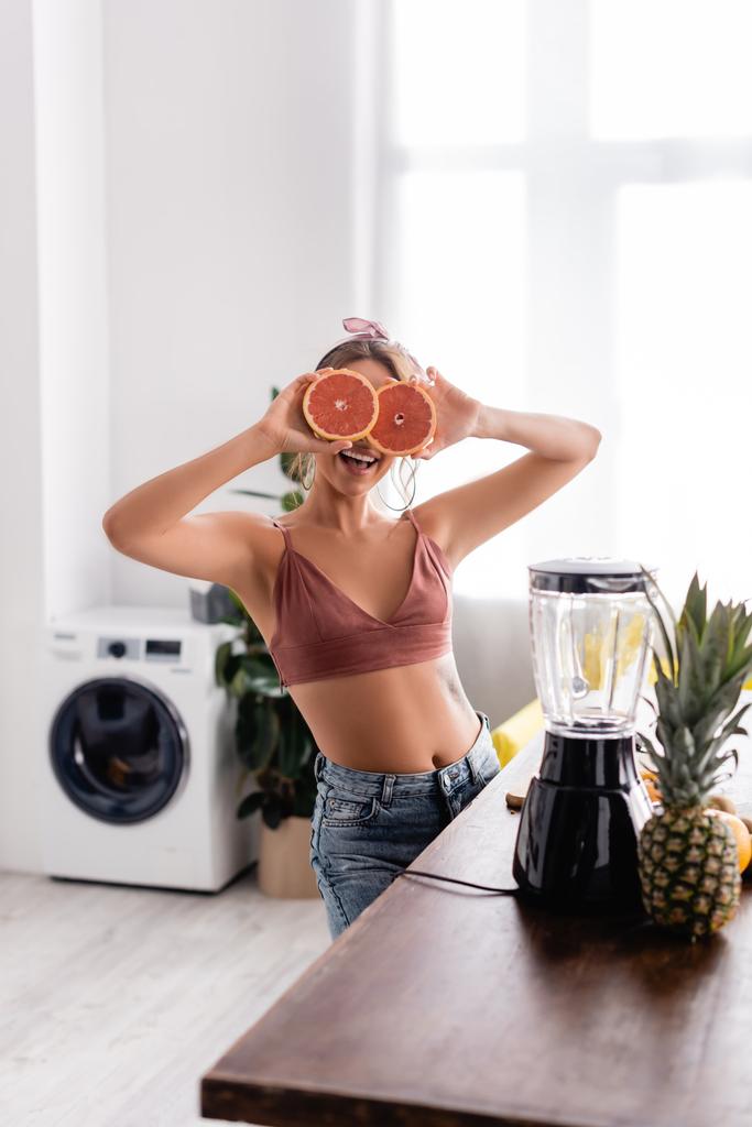 Selective focus of woman covering face with halves of grapefruit near blender on table  - Photo, Image