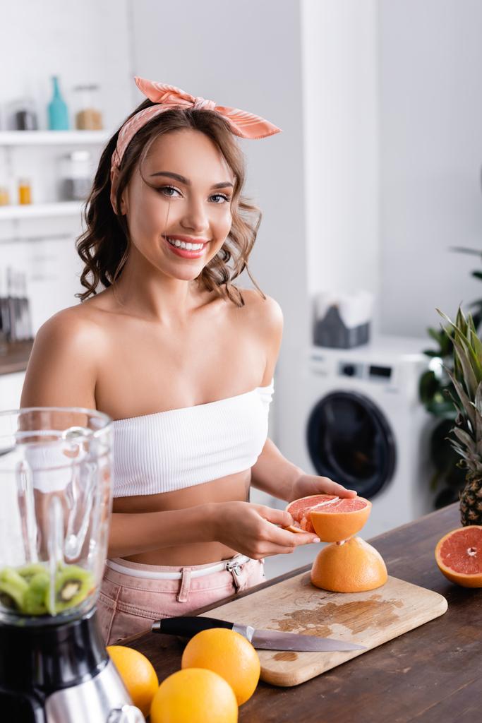 Селективное внимание домохозяйки, держащей половину грейпфрута рядом с фруктами и блендером дома  - Фото, изображение
