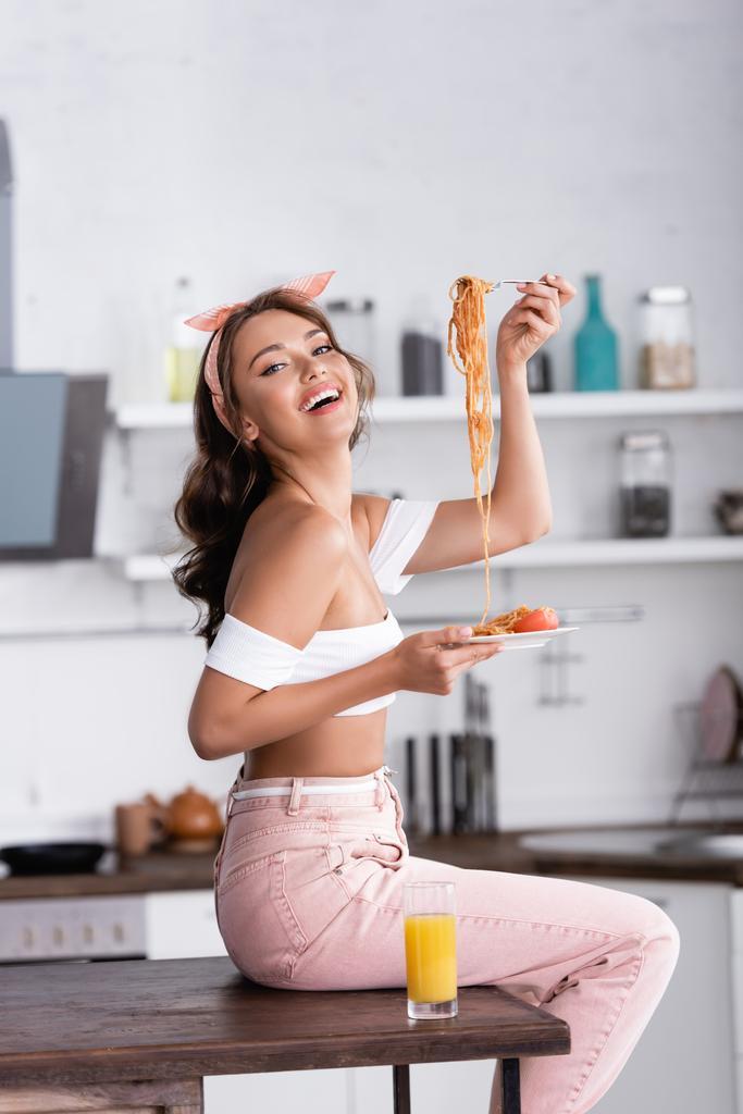 Vista lateral de la joven mujer sosteniendo espaguetis mientras está sentada en la mesa de la cocina  - Foto, imagen