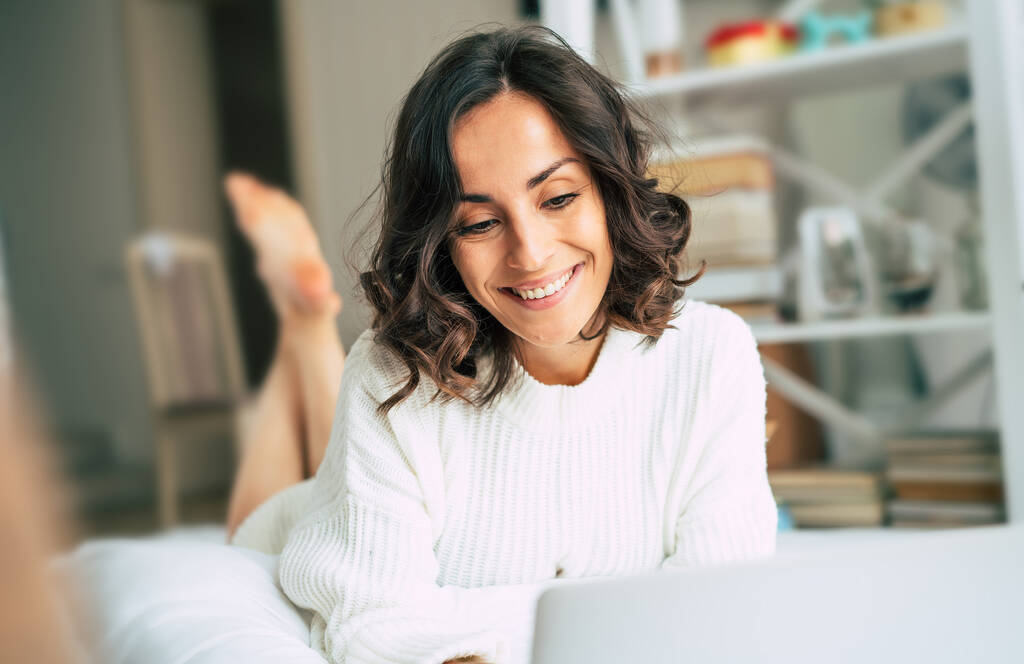 Onnellinen kaunis nuori brunette nainen työskentelee hänen laptop kun hän makasi sängyssä kotona. Freelance tyttö surffaa internetissä - Valokuva, kuva