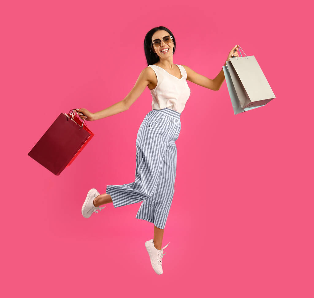 Красивая молодая женщина с бумажными пакетами покупок прыгать на розовом фоне - Фото, изображение