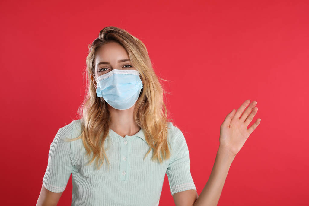 Frau mit Schutzmaske zeigt Hallo-Geste auf rotem Hintergrund. Soziale Distanz bei Coronavirus-Pandemie wahren - Foto, Bild