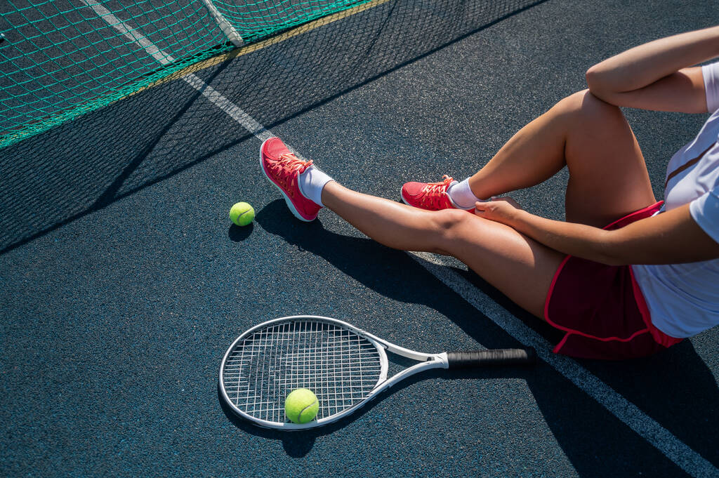 Ένα απρόσωπο κορίτσι με αθλητική φούστα κάθεται σε ένα γήπεδο τένις και κρατάει έναν πύραυλο. Κάτοψη των γυναικείων ποδιών. - Φωτογραφία, εικόνα