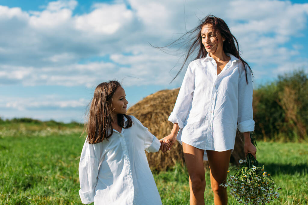Mamma e figlia, vestite allo stesso modo, con camicie bianche e fiori, passeggiano nei campi in estate. intorno al sole, verde, campo, erba, fieno, cielo. - Foto, immagini