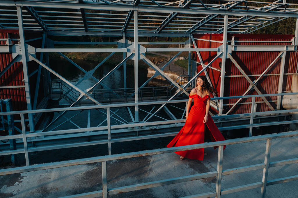長い赤のドレスで、長い茶色の髪を持つ若い美しい少女の肖像画は、今年の時間。女の子が橋の上を歩く. - 写真・画像